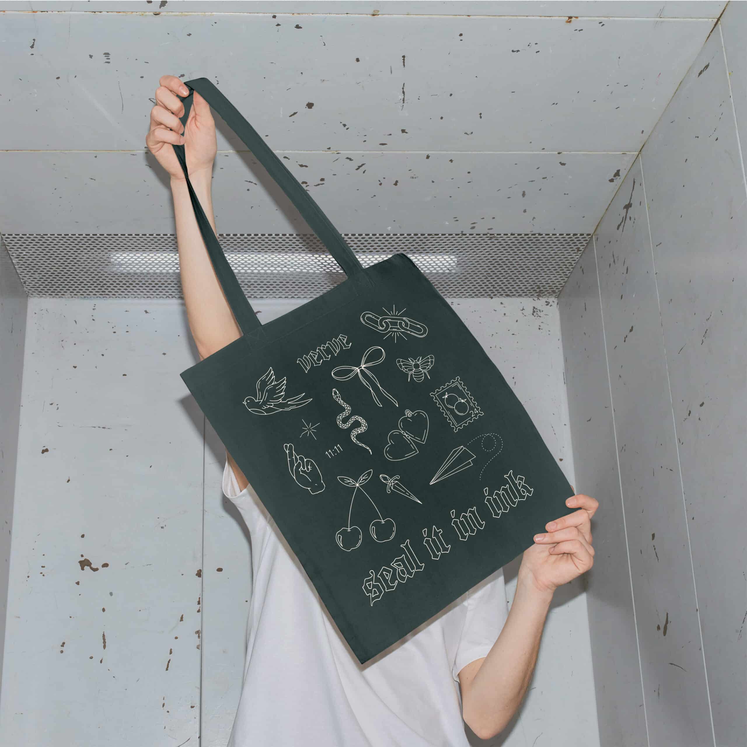 Person holding up a black tote bag with various white illustrations including cherries, a cigarette, a dagger, and text that reads "wear" and "seal it in ink.