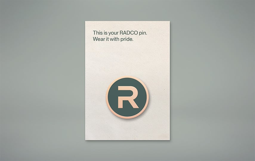 A card with a RADCO pin featuring a stylized "R" logo. The text reads, "This is your RADCO pin. Wear it with pride.