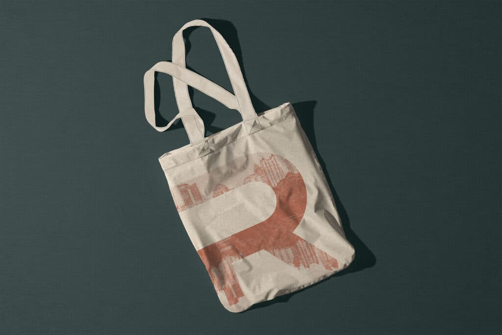 A crumpled white tote bag with two handles on a dark surface. The bag features a large, abstract, rust-colored letter "R" design.