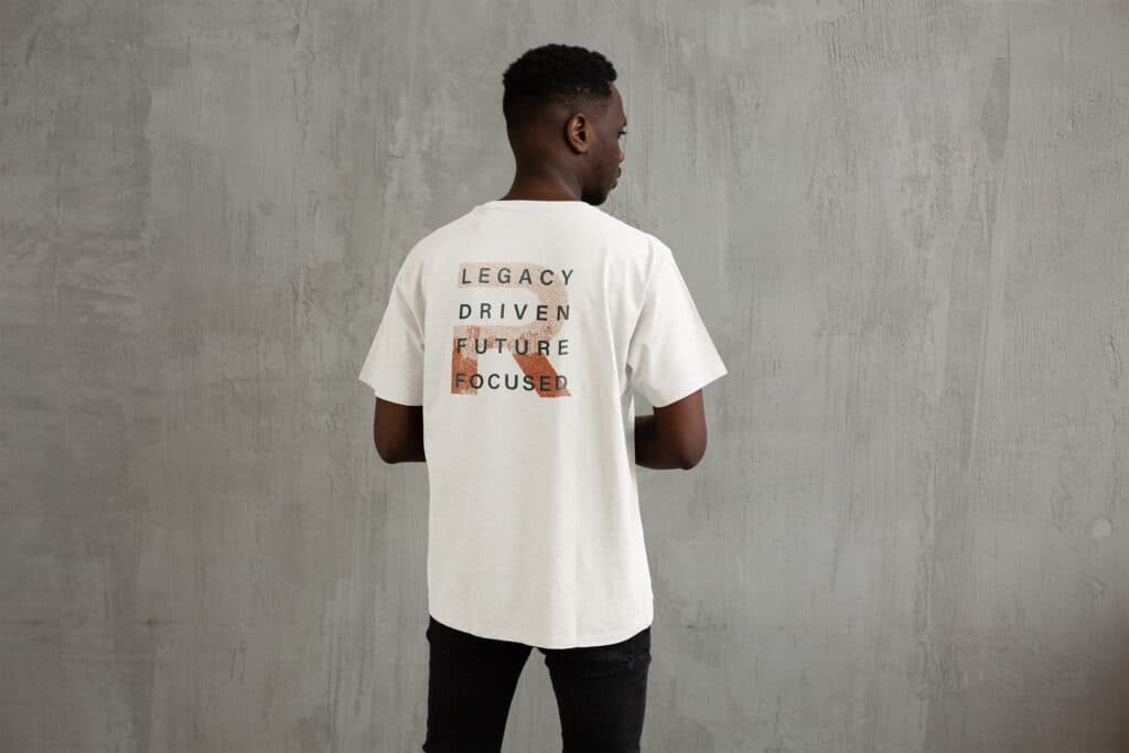 Man wearing a white t-shirt with the text "Legacy Driven Future Focused" stands against a gray textured wall.