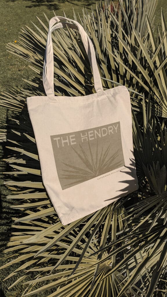 Tote bag with "The Hendry" design lies on a palm-like plant in a grassy area.