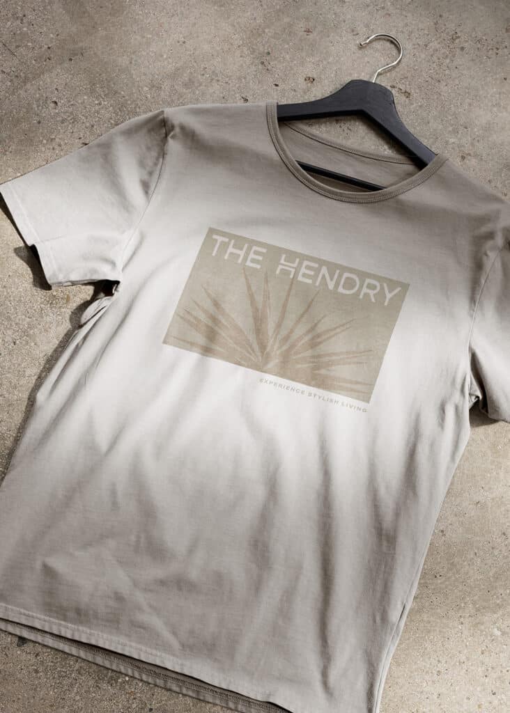 Beige T-shirt on a hanger with a graphic design and text "THE HENDRY, EXPERIENCE STYLISH LIVING.